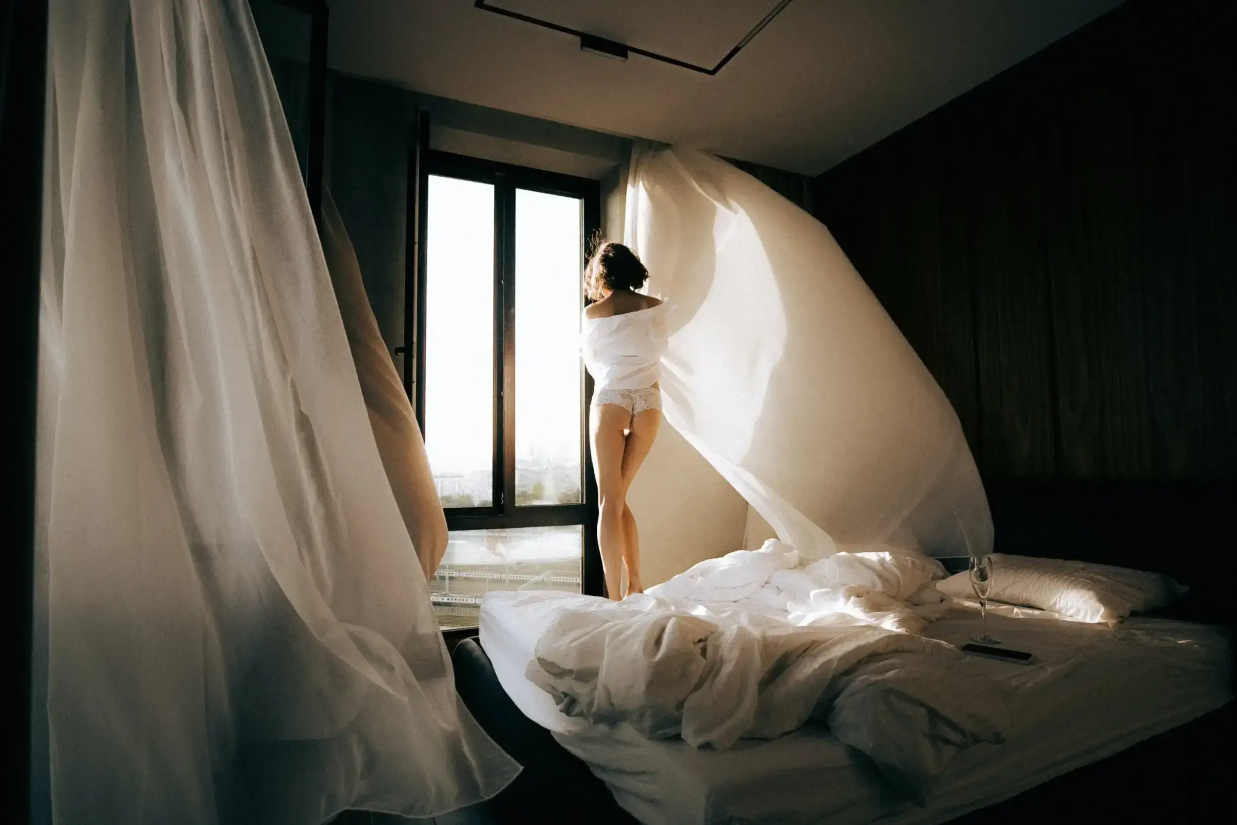 Woman standing by bedroom window with billowing white curtains.